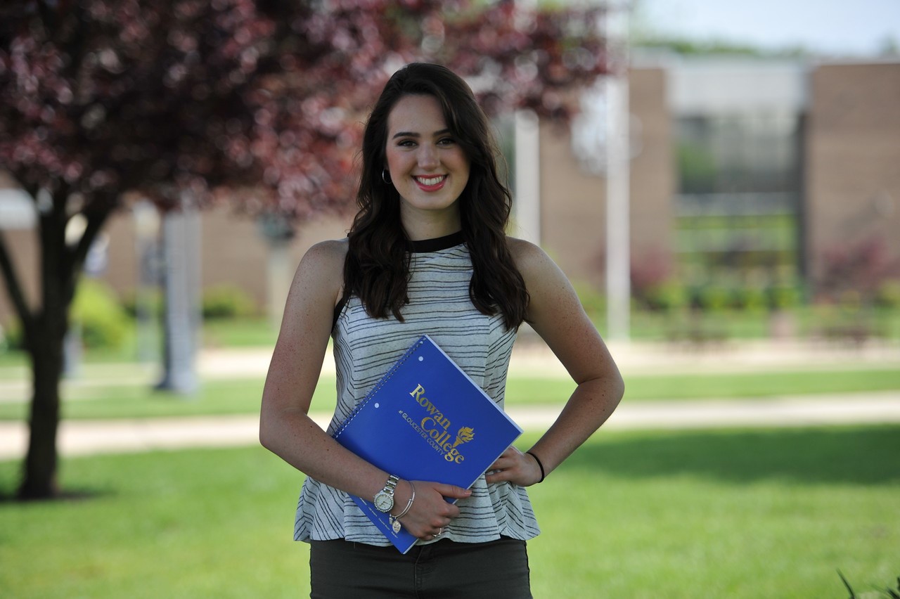 A photo of a student smiling standing and holding a book on the Gloucester Campus.